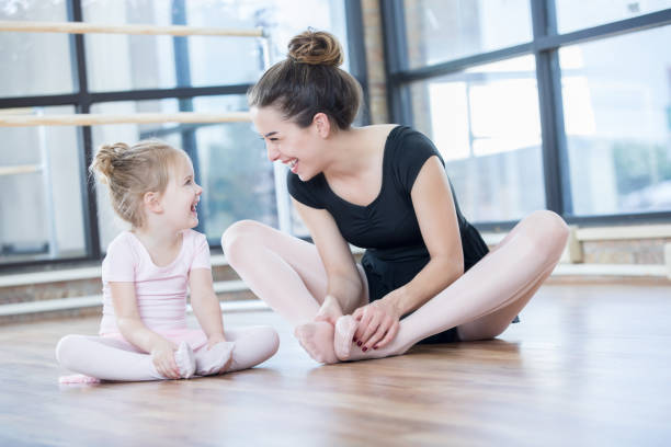 A young ballet instructor sits on the floor with her adorable young preschool age student as they look at each other and laugh while stretching.